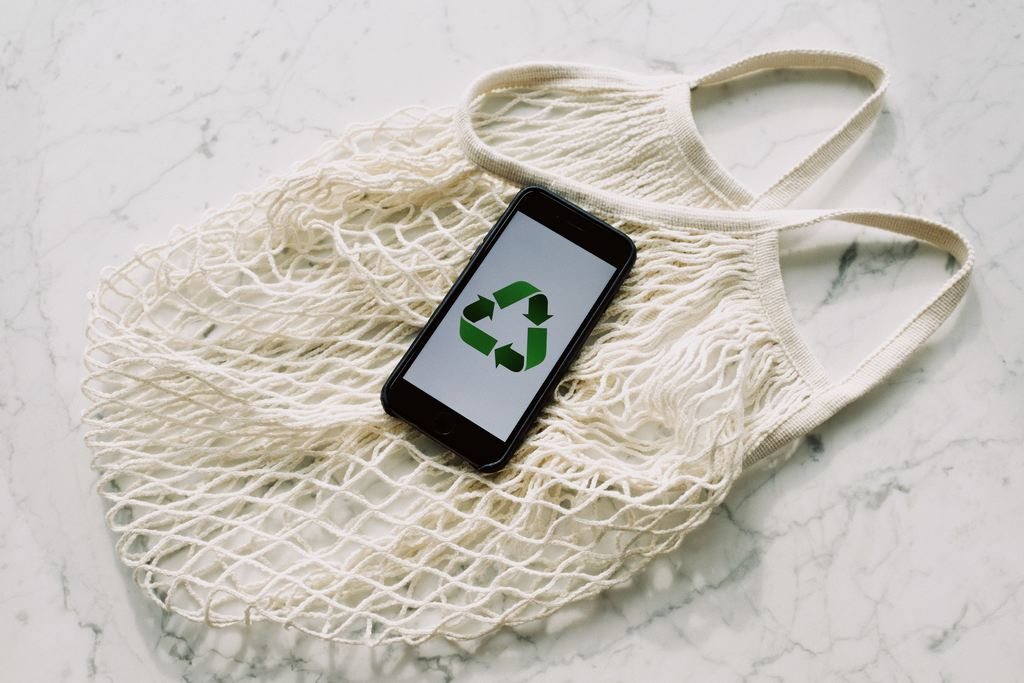 Um celular cuja tela mostra o símbolo de reciclável. O celular está sobre uma sacola reutilizável (eco bag), simbolizando embalagens eco-friendly