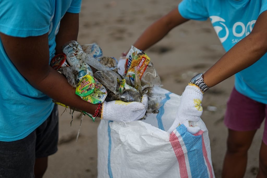 Uma pessoa abrindo um saco para outra jogar resíduos recicláveis dentro, como embalagens amassadas. Simbolizando a importância da reciclagem no Brasil