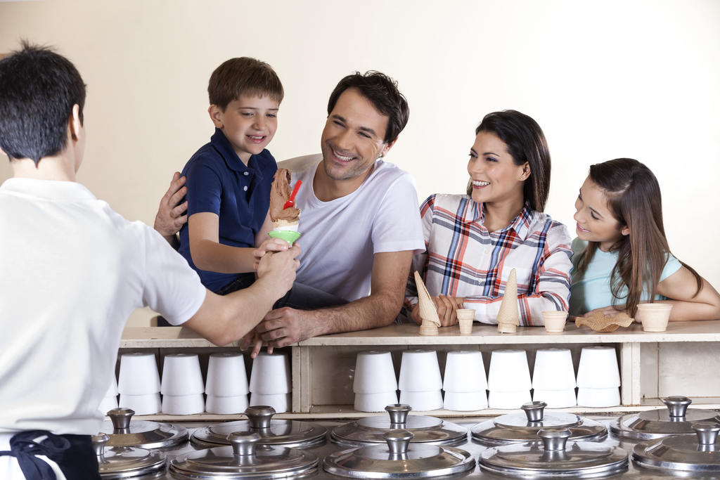 Família (um casal de adultos e um casal de crianças) dentro de uma sorveteria, sorridentes. O menino pequeno está pegando um sorvete de massa com casquinha que o sorveteiro está entregando para ele.