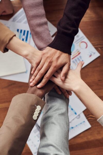 Grupo com as mãos juntas, num acordo de motivação, simbolizando Responsabilidade social corporativa. Ao fundo, sobre uma mesa, estão documentos, gráficos, um caderno aberto e um notebook.