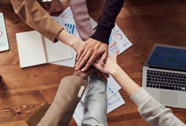 Grupo com as mãos juntas, num acordo de motivação, simbolizando Responsabilidade social corporativa. Ao fundo, sobre uma mesa, estão documentos, gráficos, um caderno aberto e um notebook.