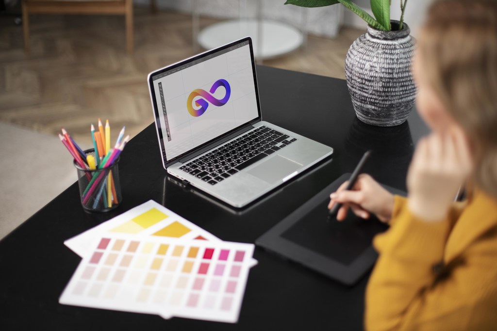 Mulher criando um logotipo no notebook usando mega digitalizadora para desenhar. Ao lado do notebook existem duas folhas de papel com paletas de cores e um pote com lápis de cor. O logotipo representa fortalecimento de marca