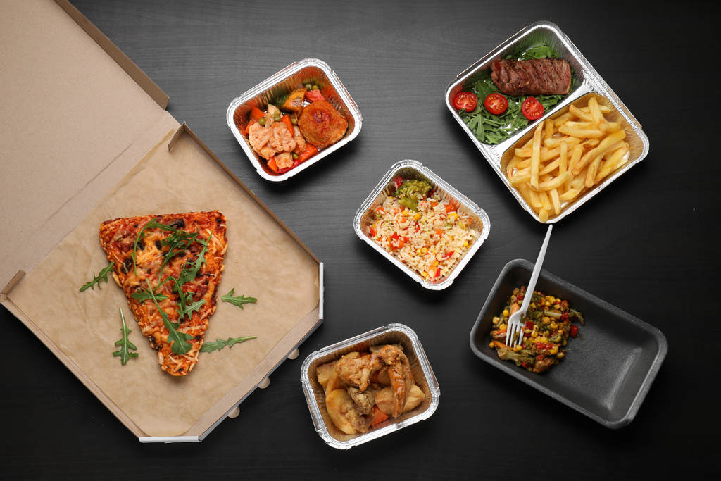 Diferentes tipos de embalagens para delivery, como embalagem de papel com pizza dentro, embalagens de marmita de alumínio com arroz, frango, carne com salada e tomate, batata frita e outros alimentos. Além de uma embalagem de marmita de isopor preto com garfinho de plástico.