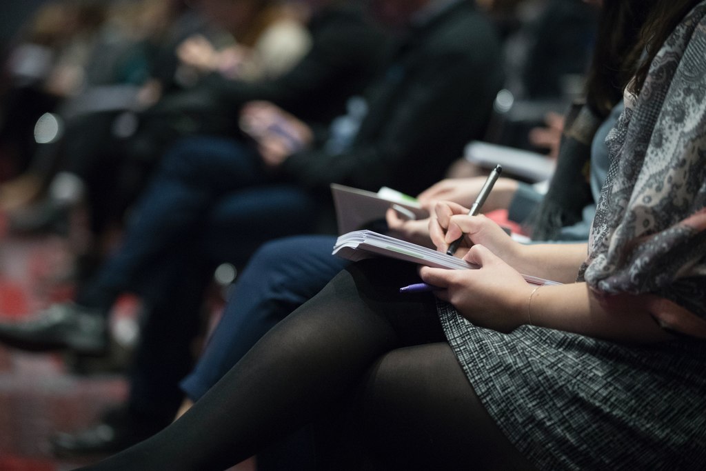 Uma espécie de congresso com pessoas na plateia fazendo anotações, simbolizando treinamentos sobre práticas sustentáveis nas empresa