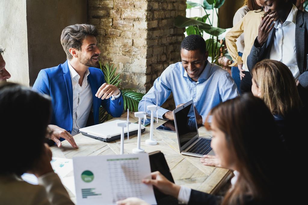 Grupo de pessoas sorrindo, de roupa social, sentadas à uma mesa de reunião com notebooks e documentos com gráficos, representando exemplos de práticas sustentáveis nas empresas