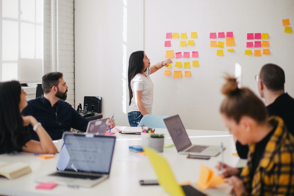 Mulher fazendo reunião usando técnicas para marketinh de eventos corporativos num quadro branco com post its, dentro de uma sala com mais quatro colaboradores que estão sentados à mesa de reunião, a qual trem notebooks, canetas e blocos de anotações