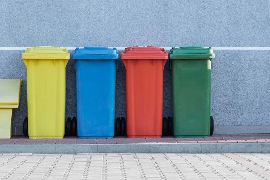 Cestos de lixo grandes, de diferentes cores (amarelo, verde, vermelho e verde). Simbolizando coleta seletiva de materiais recicláveis