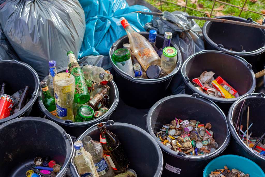 Materiais recicláveis e não recicláveis, separados em sacos de lixo e em baldes abertos. Nos baldes: tipos de lixo recicláveis separados em baldes, como de vidro (garrafas e potes), tampinhas de alumínio, latinhas e varetas de plástico.