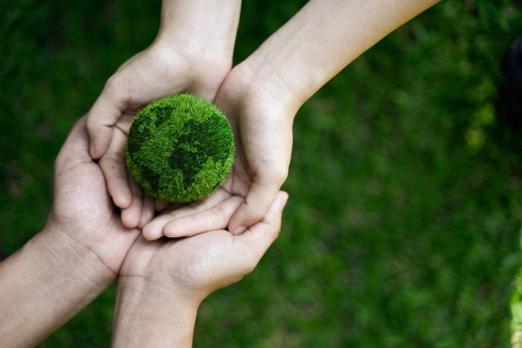 Mãos de adulto sob mão de criança, que está segurando um globo terrestre feito de vegetação verde, simbolizando carbono zero