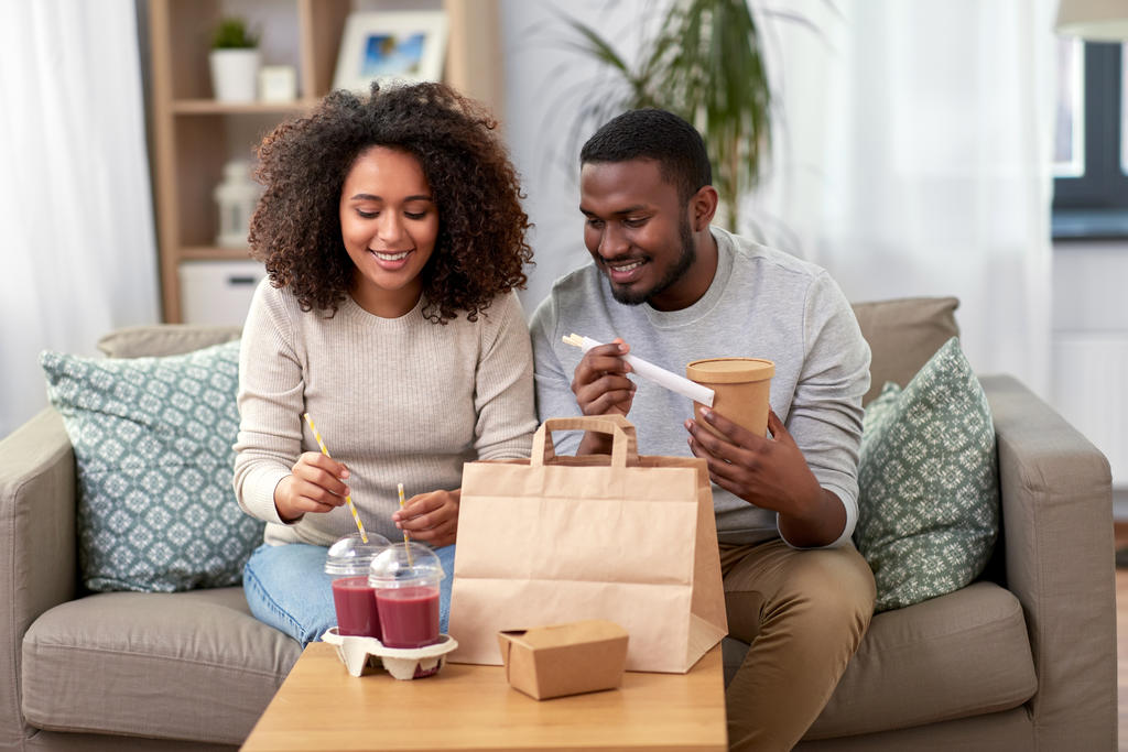 Casal na sala de estar, sorrindo olhando para delivery sustentável que está sobre a mesa de centro. A mulher está colocando canudos sustentáveis em dois copos com tampa. Há uma sacola de papel sobre a mesa e uma caixa para marmita. O homem está segurando um pote sustentável e hashis ou talheres de madeira.