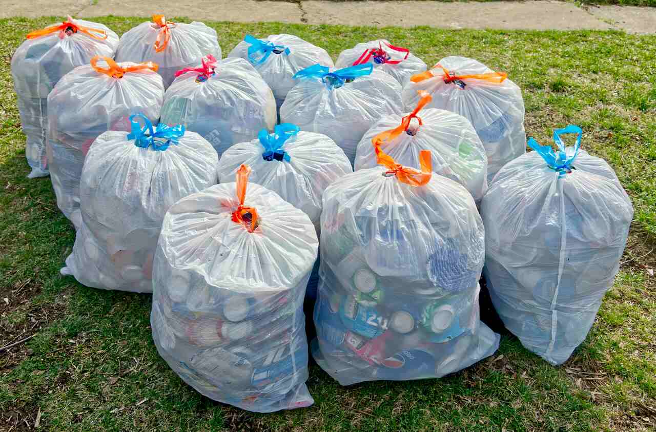 Vários sacos transparentes cheios de materiais recicláveis organizados sobre a grama, com fitas coloridas fechando cada saco.