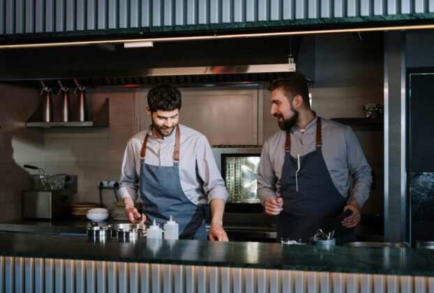 Dois cozinheiros trabalhando lado a lado em uma cozinha profissional, usando aventais e preparando alimentos na bancada.