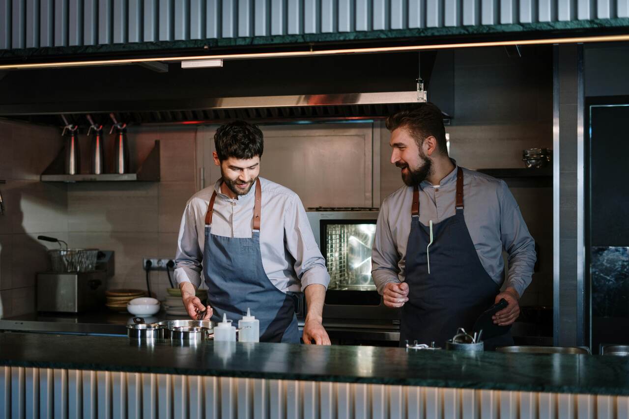 Dois cozinheiros trabalhando lado a lado em uma cozinha profissional, usando aventais e preparando alimentos na bancada.