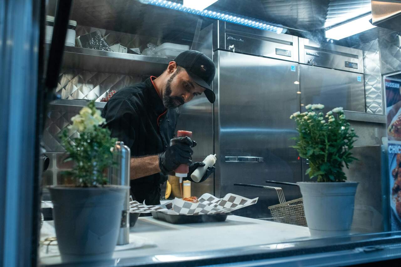 Cozinheiro em food truck aplicando molho em um sanduíche, com prateleiras de inox e plantas ao redor.