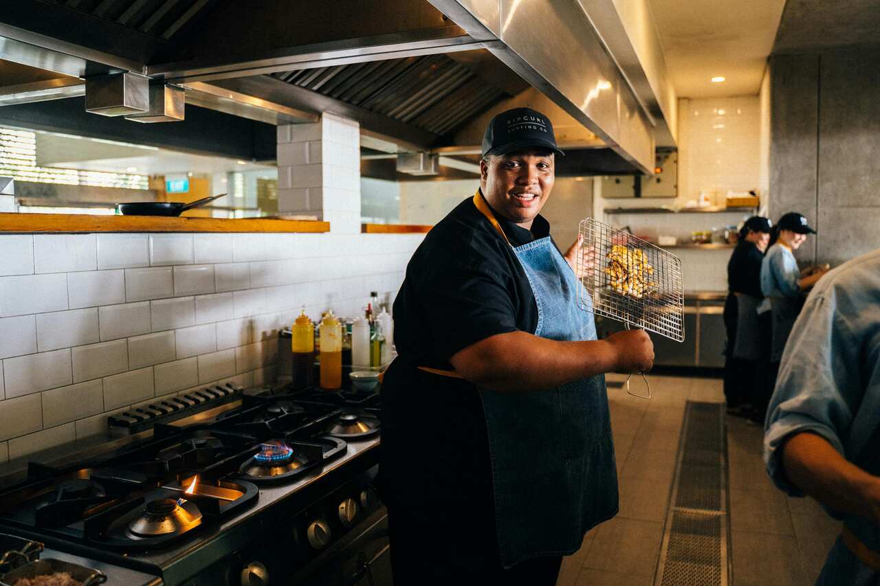 Cozinheiro sorridente segurando uma grelha com alimentos assados em uma cozinha industrial, com fogões e utensílios ao fundo.
