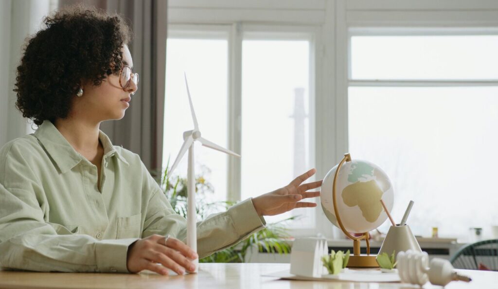 Mulher observando um globo terrestre enquanto segura uma mini turbina eólica sobre a mesa, em ambiente iluminado.