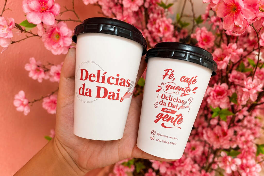 Pessoa segurando copos para delivery personalizados para café quente com tampa e identidade visual da marca em fundo de flores
