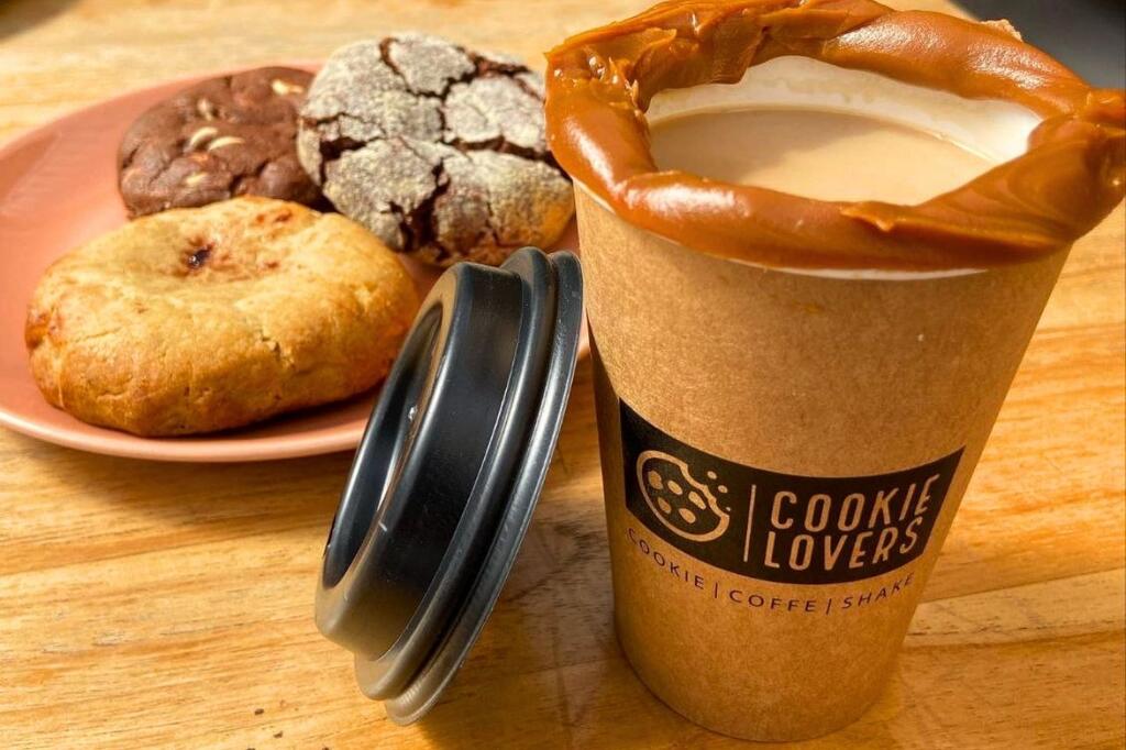 Copo personalizado para cafeteria com tampa ao lado de um prato de cookies em cima de uma mesa